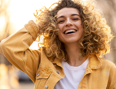 IMAGE: Young woman in golden jacket laughing with her hand in her blond curly hair.