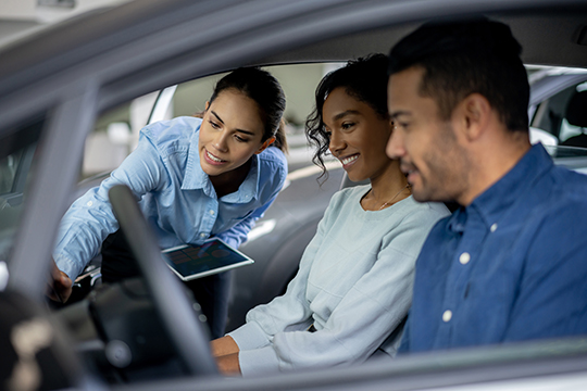 IMAGE: Couple sitting inside car at dealership and salesperson showing the features