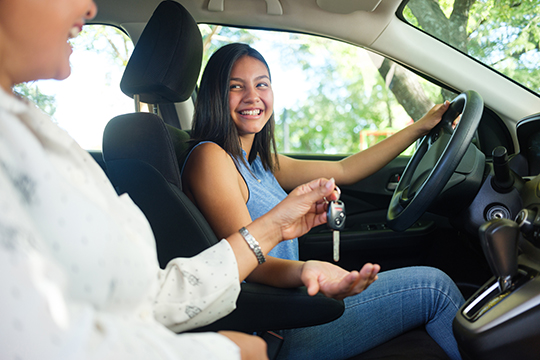 IMAGE: Female teen driver smiling behind wheel and being handed keys to the vehicle.