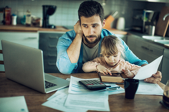 IMAGE: Worried man with young daughter in his lap at a table with wallet, papers, and a calculator
