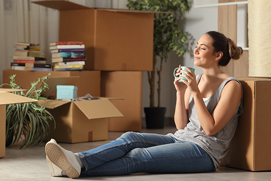 IMAGE: Woman sitting on floor with coffee mug smiling and moving boxes open around