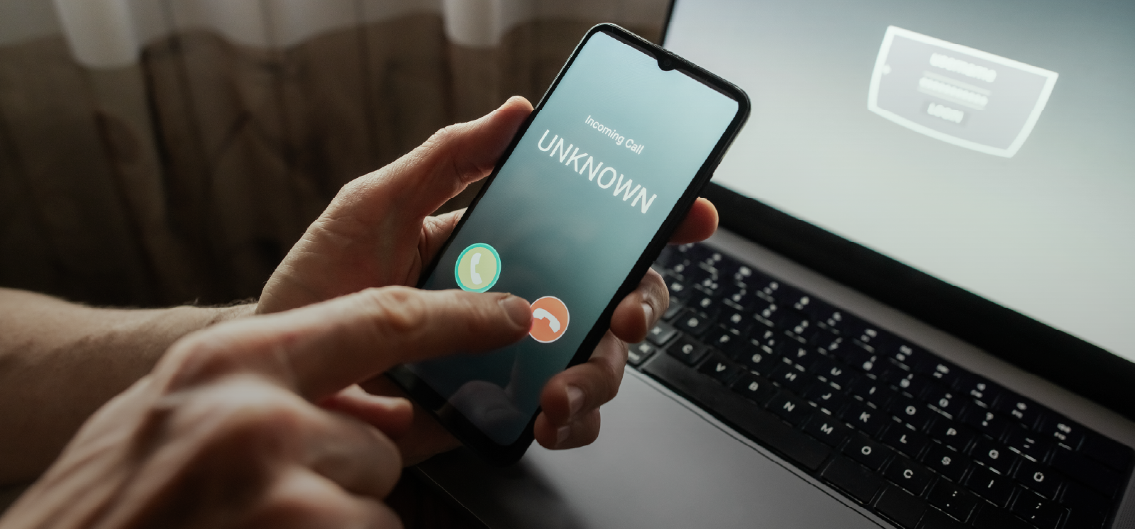 IMAGE: Person blocking unknown phone number on cell phone with laptop open in background