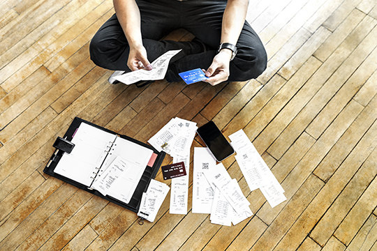 IMAGE: Person sitting on floor holding credit card and receipts with more receipts, cards, and a planner scattered on floor
