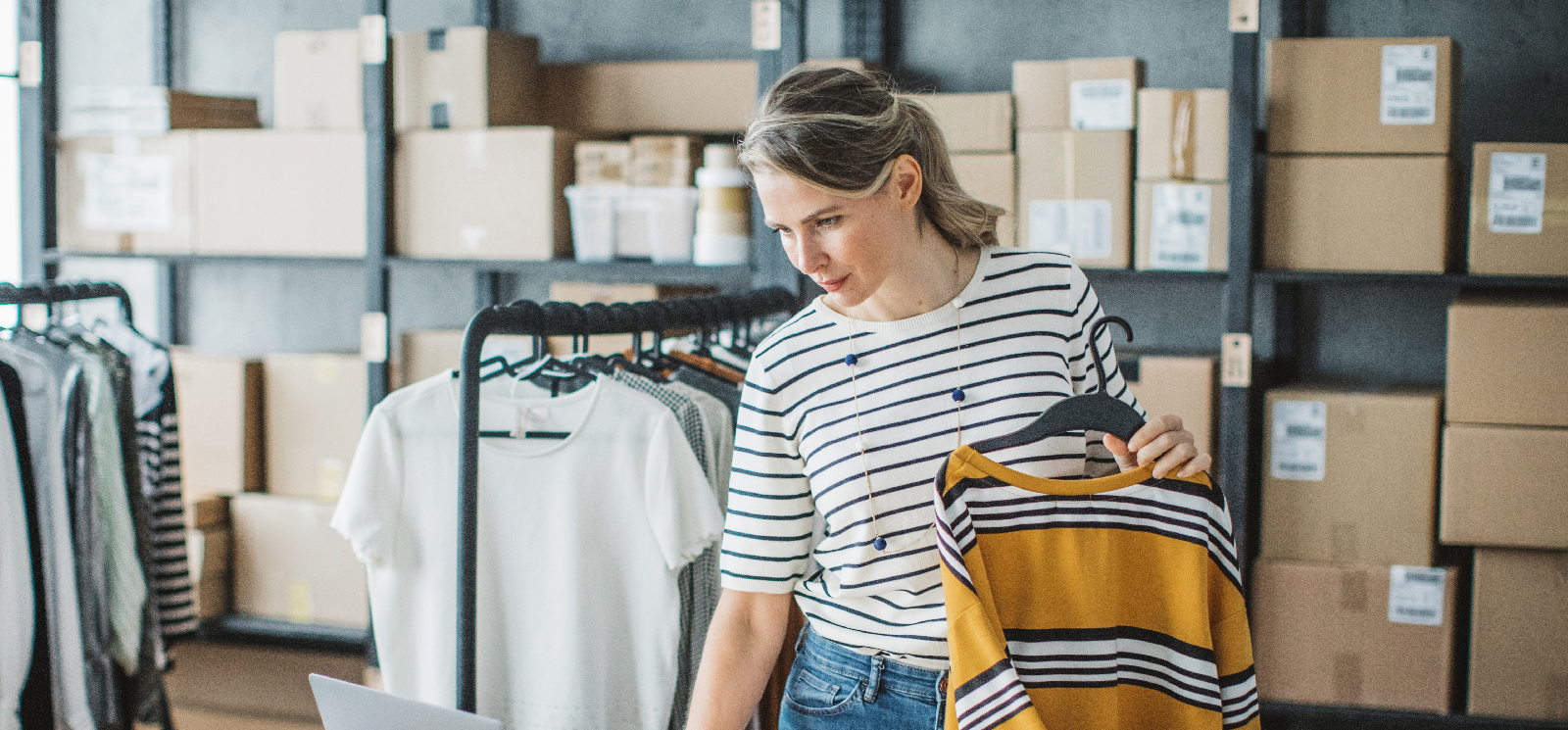 IMAGE: Woman holding shirt while looking at a laptop inside a storage area for a clothing store